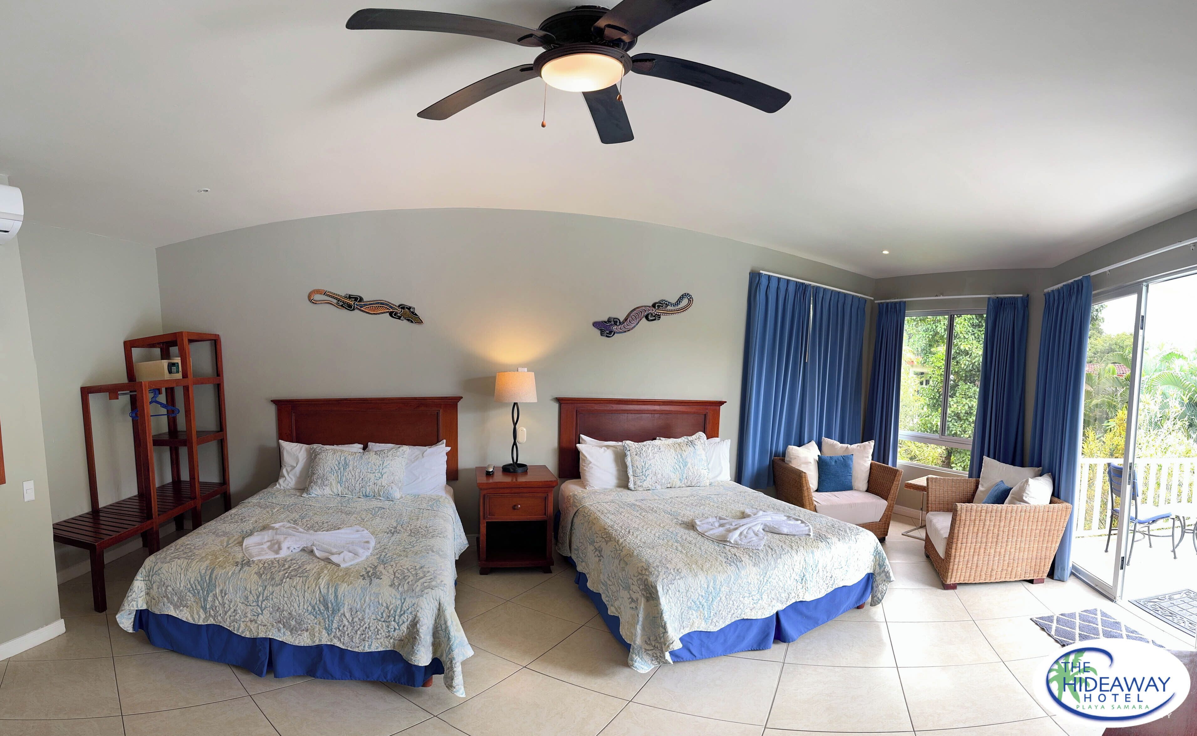 A hotel room with two beds, a ceiling fan, a nightstand, a lamp, a decorative wall hanging, and a view of the outdoors.