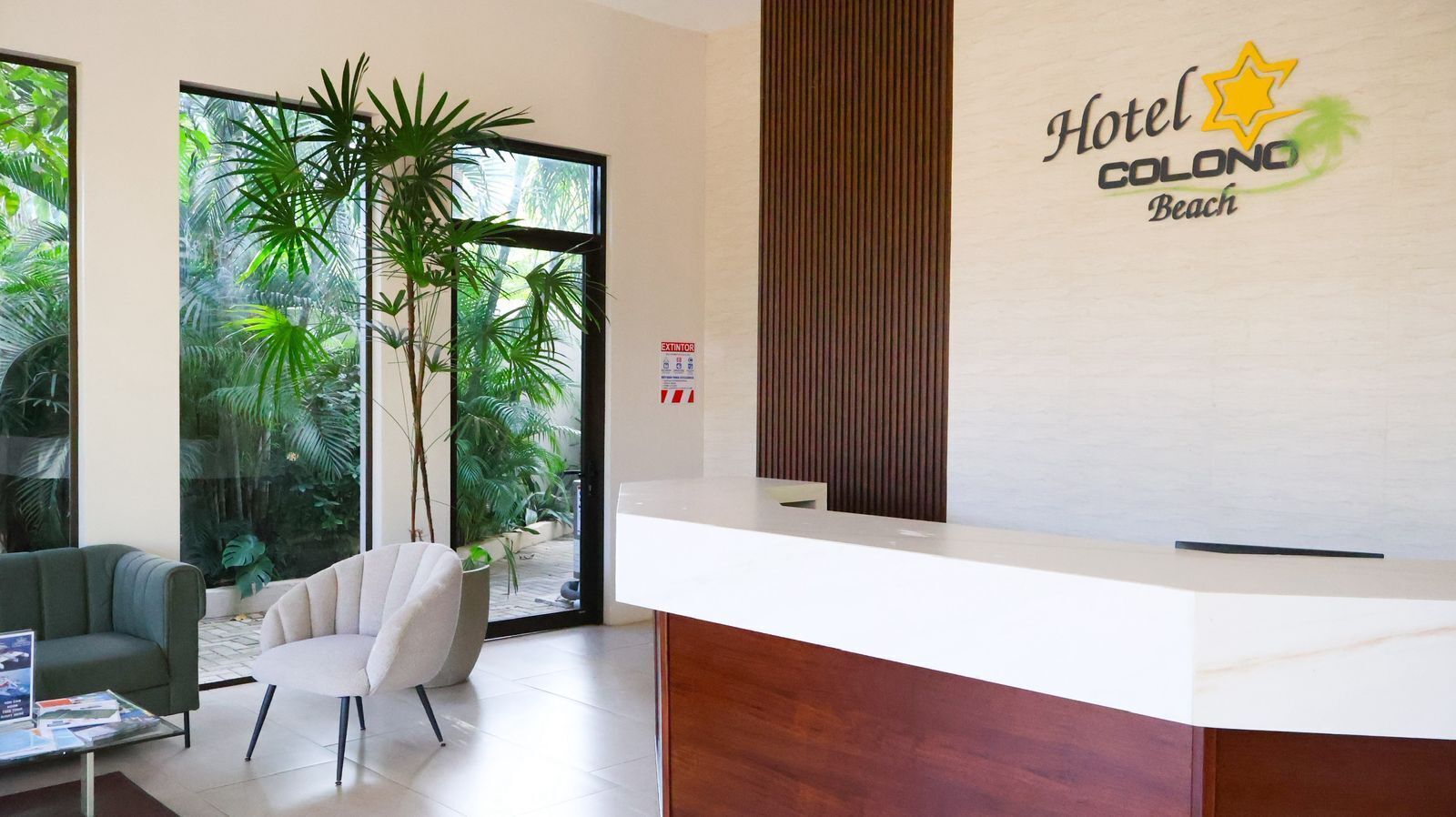 A hotel reception area with a wooden reception desk, a green sofa, a white armchair, and a large window with tropical plants.
