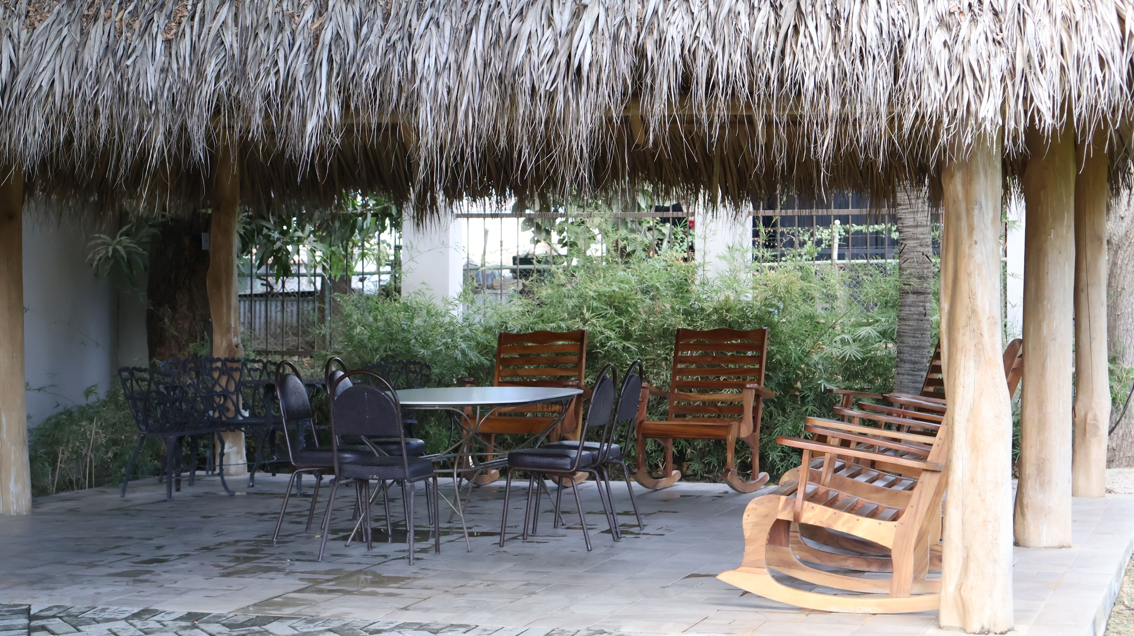 An outdoor seating area with a thatched roof, surrounded by greenery and wooden furniture.