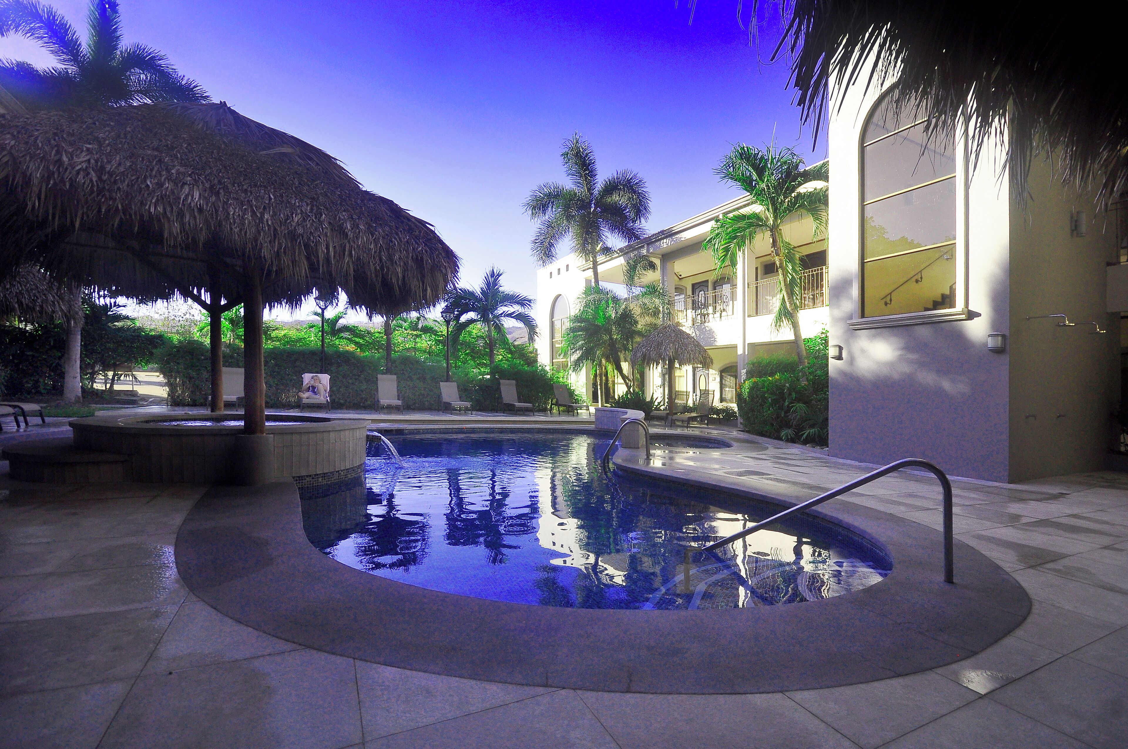 Una piscina con techo de paja, palmeras y un edificio con balcones.