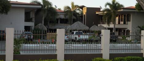 Un hotel con estacionamiento cercado, palmeras y sombrillas de paja.