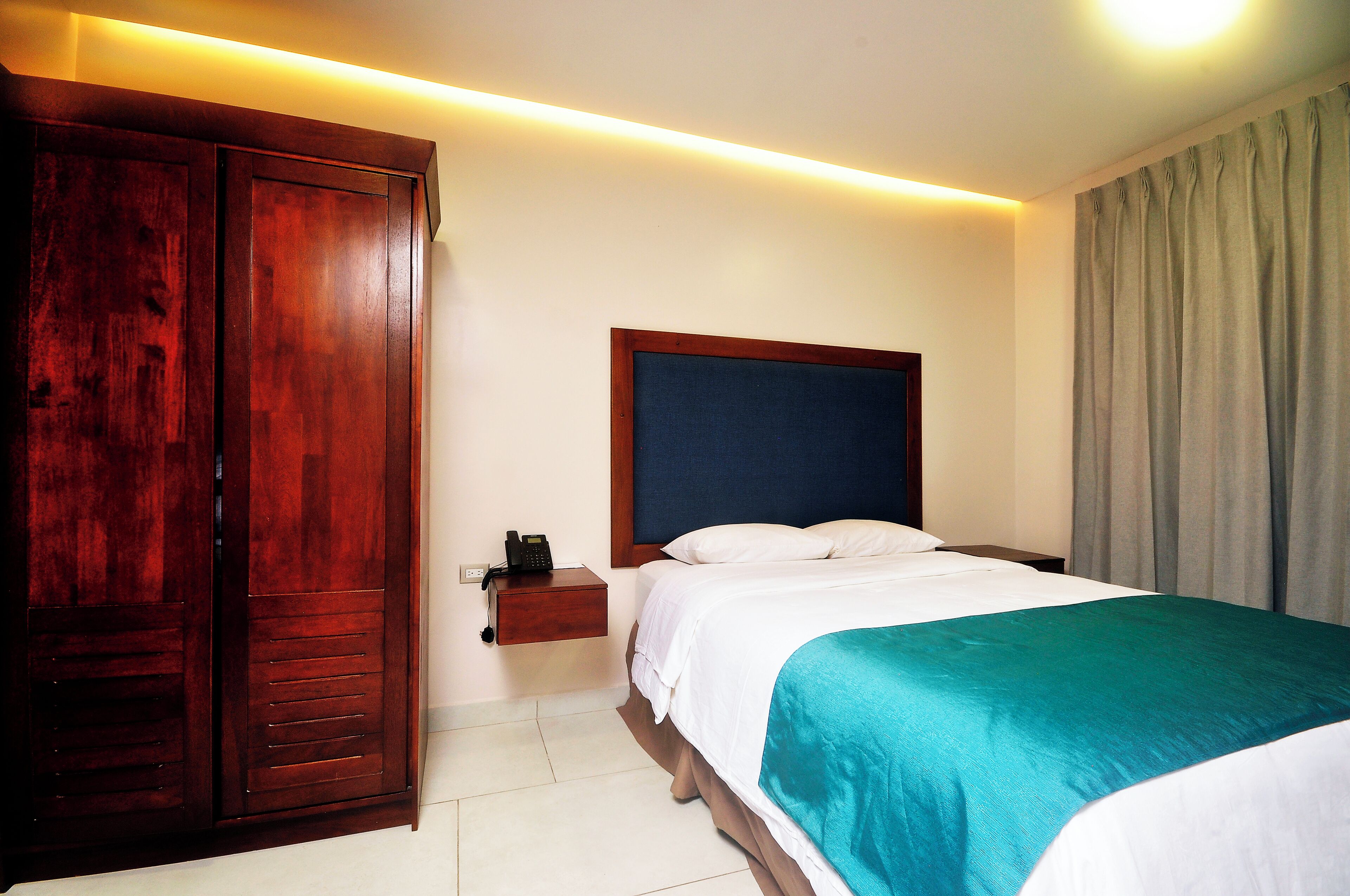 A hotel room with a wooden wardrobe, a bedside table with a phone, a bed with white and teal bedding, and a headboard.