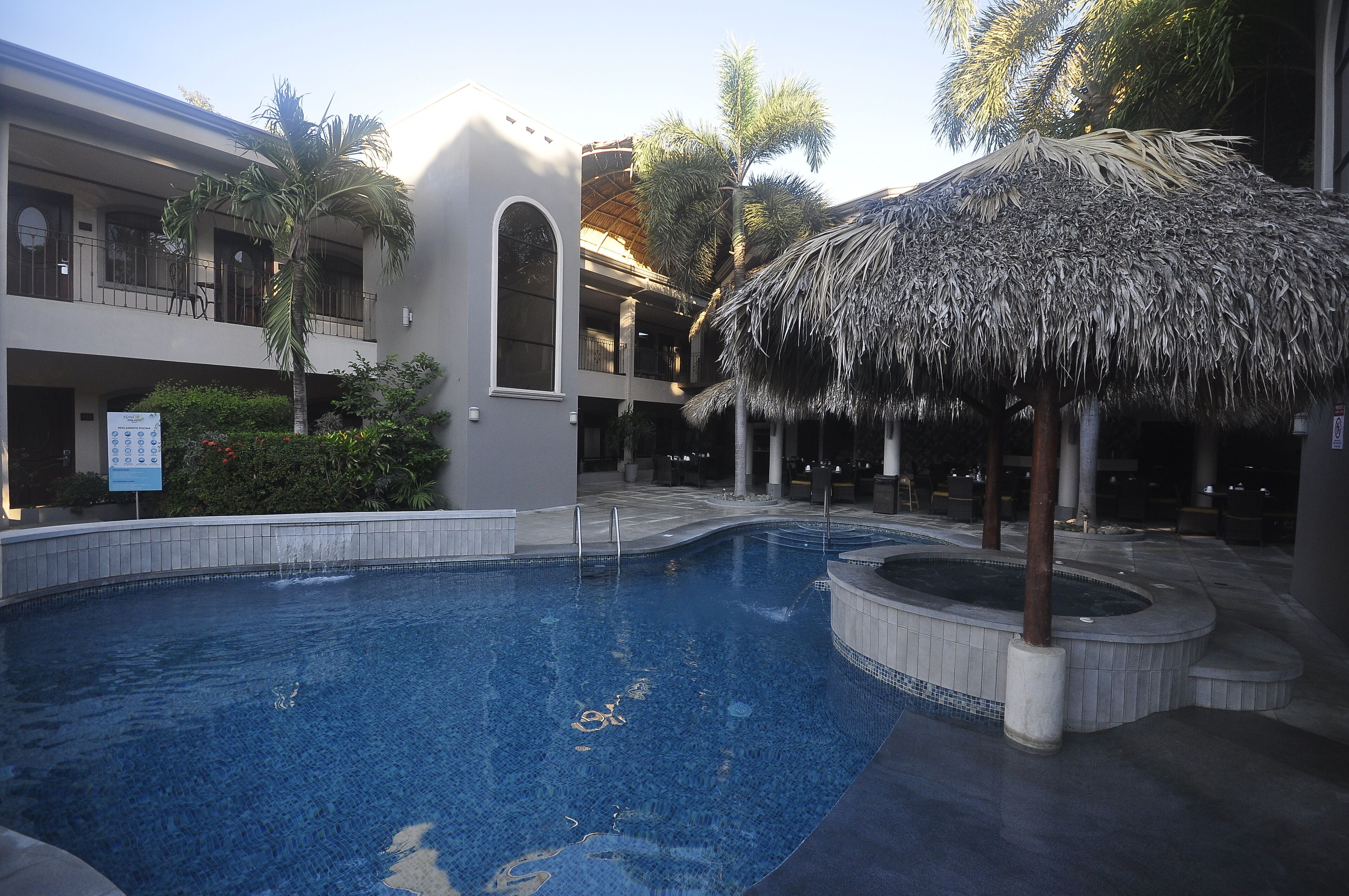 A swimming pool with a thatched roof structure, a multi-story building with balconies, and a signboard.