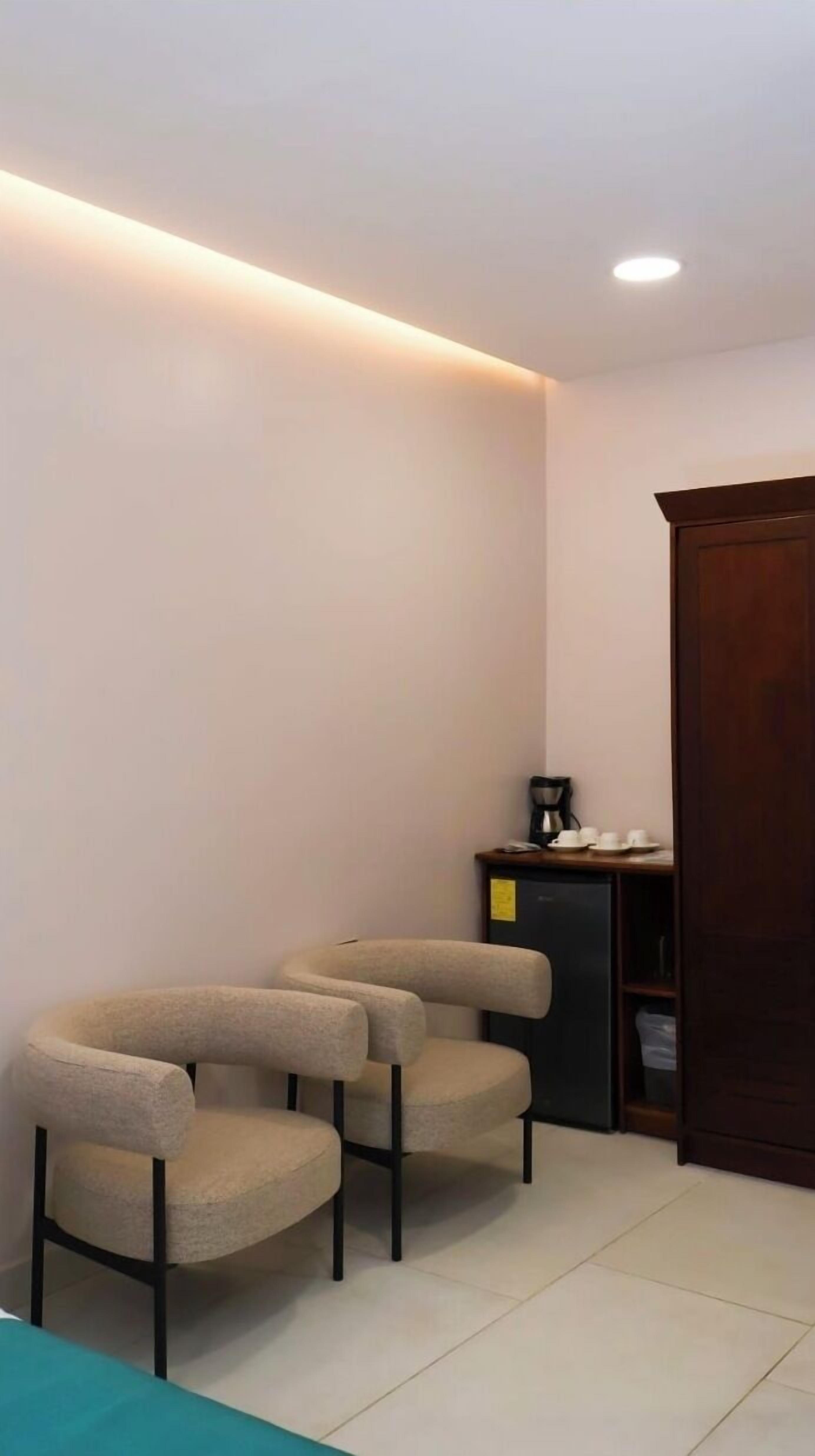 A corner room with two beige armchairs, a small table with a coffee maker, and a dark wooden cabinet.