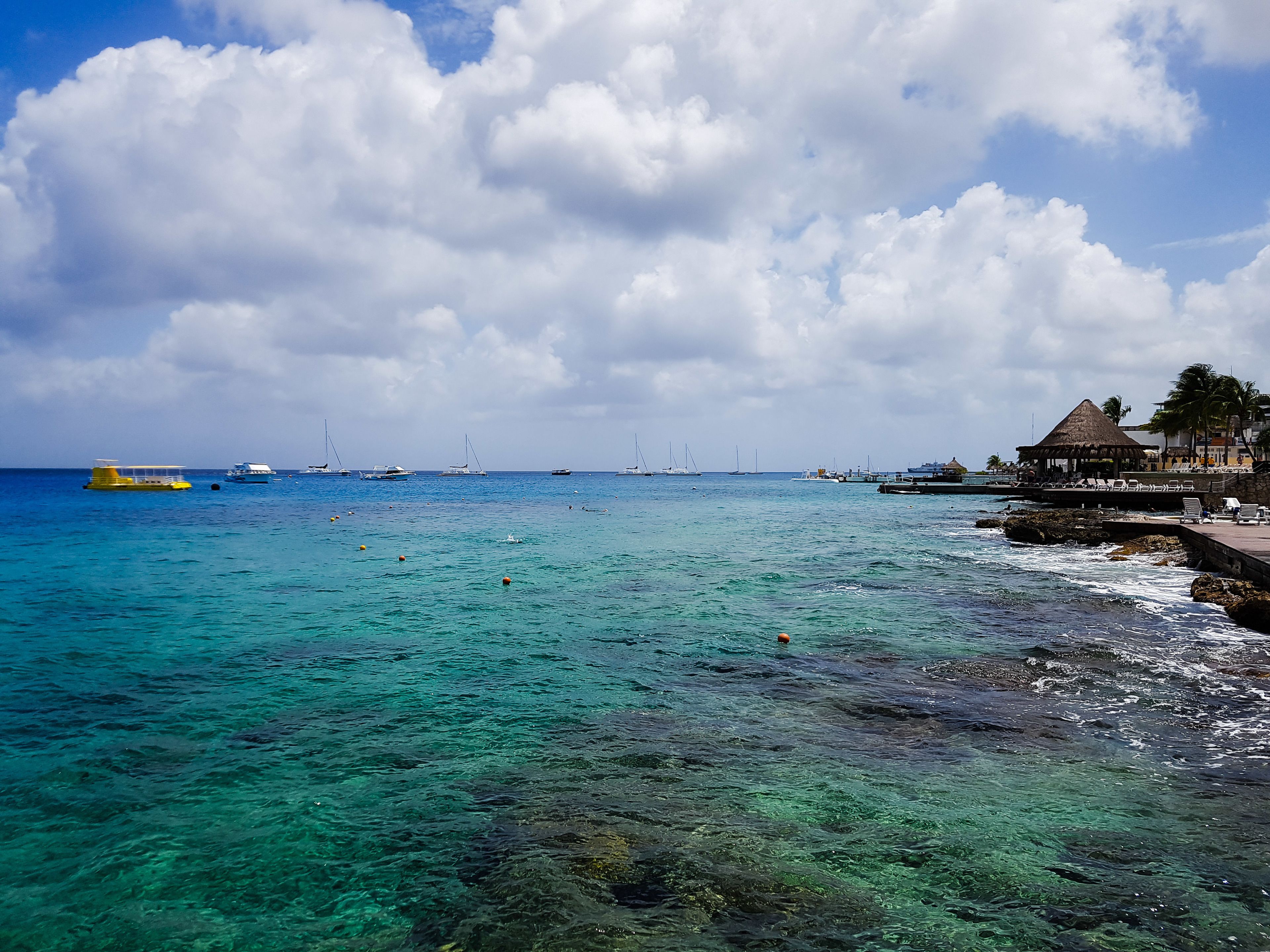A coastal scene with clear turquoise waters, boats, and a beach area with a thatched-roof structure.