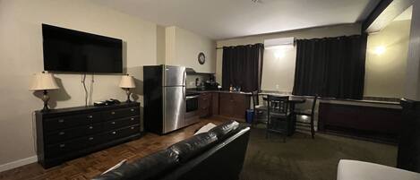 Un espacio habitable compacto con kitchenette, un televisor de pantalla plana montado en la pared, un sofá de cuero y un comedor con mesa y sillas.