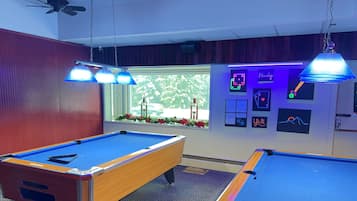 A pool table with a blue felt surface, a wooden pool table, and a window with a view of trees and a car.