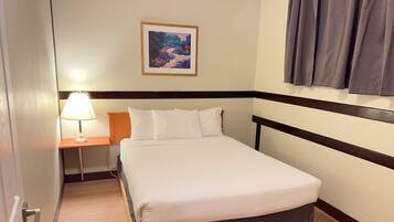 Une chambre d’hôtel avec un lit, une table de chevet avec une lampe, un tableau encadré au mur et une fenêtre avec des rideaux.