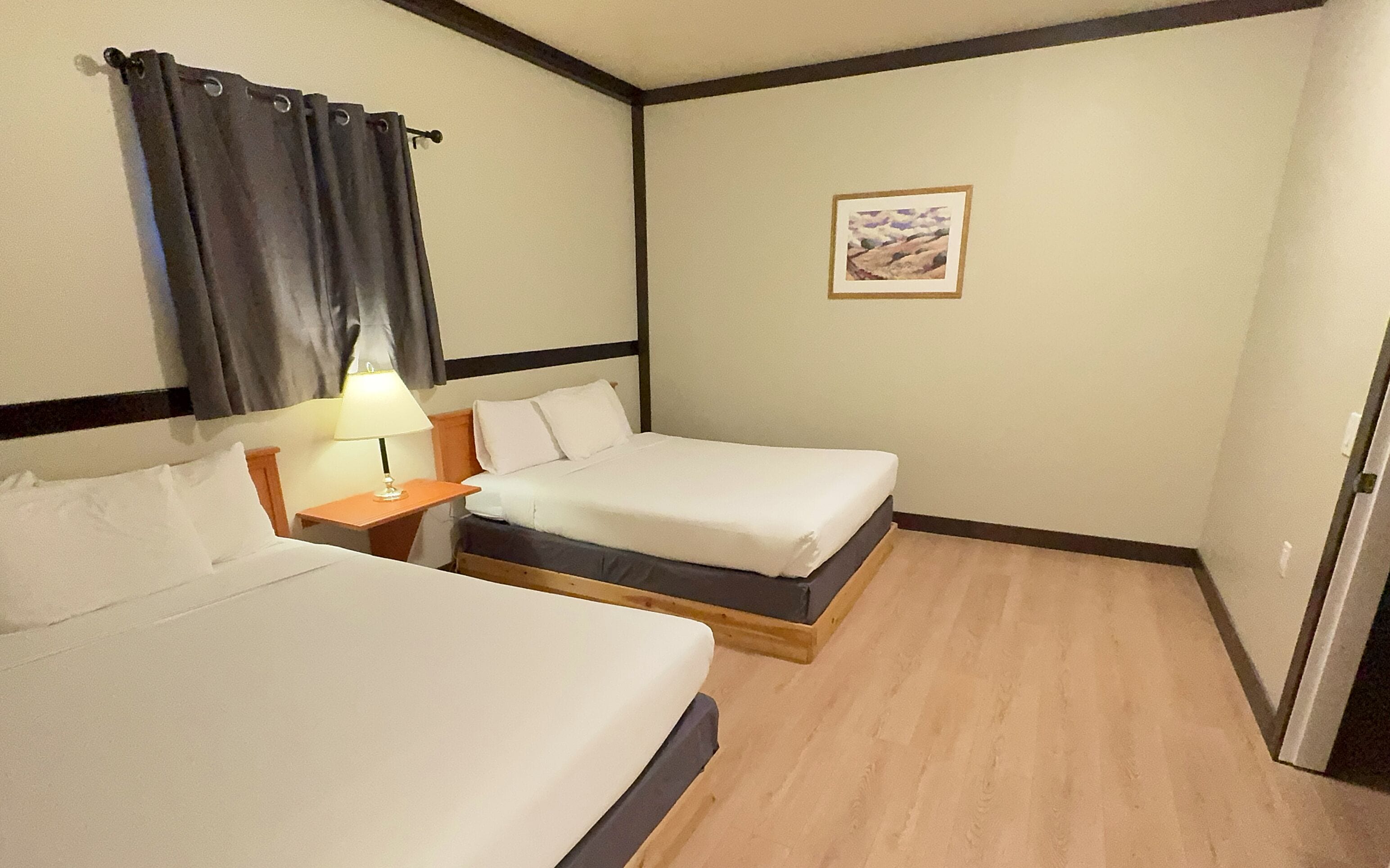 A hotel room with two single beds, a nightstand with a lamp, a framed picture on the wall, and a wooden floor.