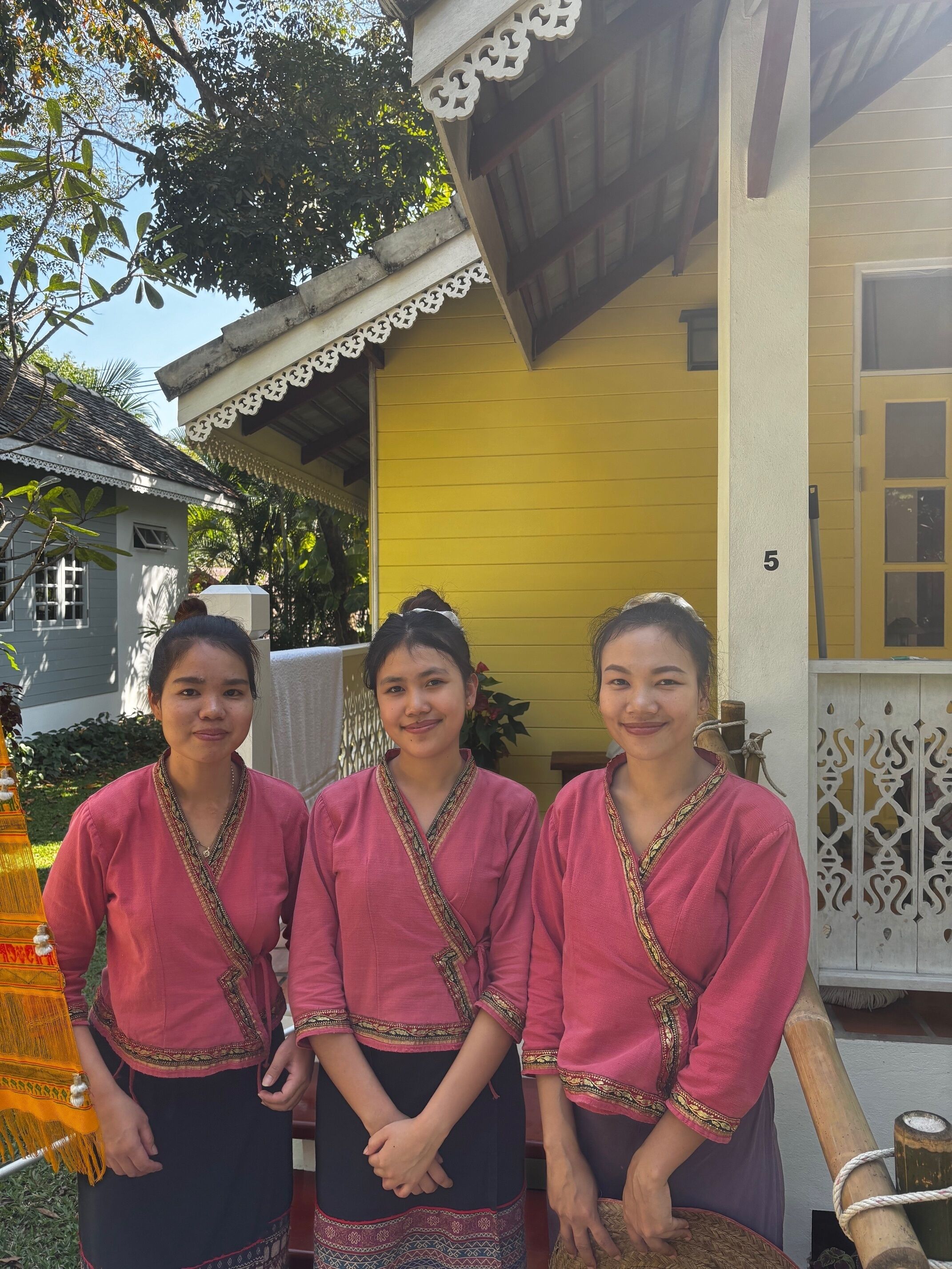Three individuals standing in front of a yellow building with a white decorative railing and a number '5' on the wall.