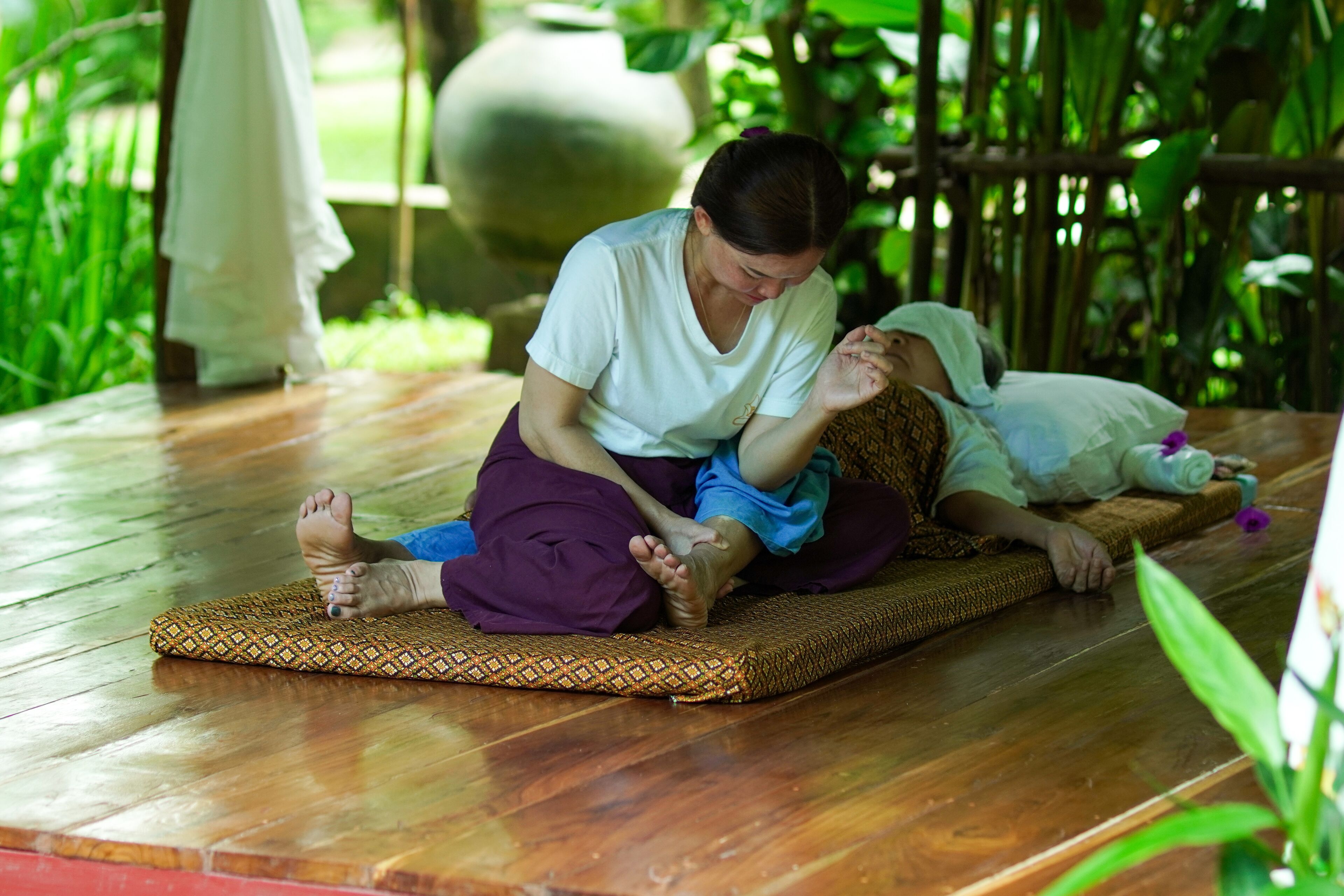 A person lying on a mat in a room with a wooden floor and a large stone in the background.