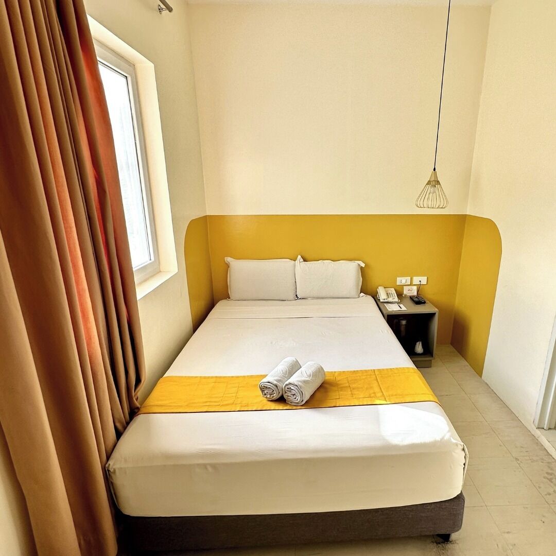 Habitación de hotel con cama, mesita de noche con teléfono y lámpara, y ventana con cortinas.