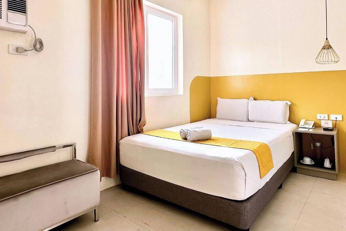 Une chambre d’hôtel comprenant un lit, une table de chevet avec un téléphone et une lampe, une fenêtre avec des rideaux et un climatiseur fixé au mur.
