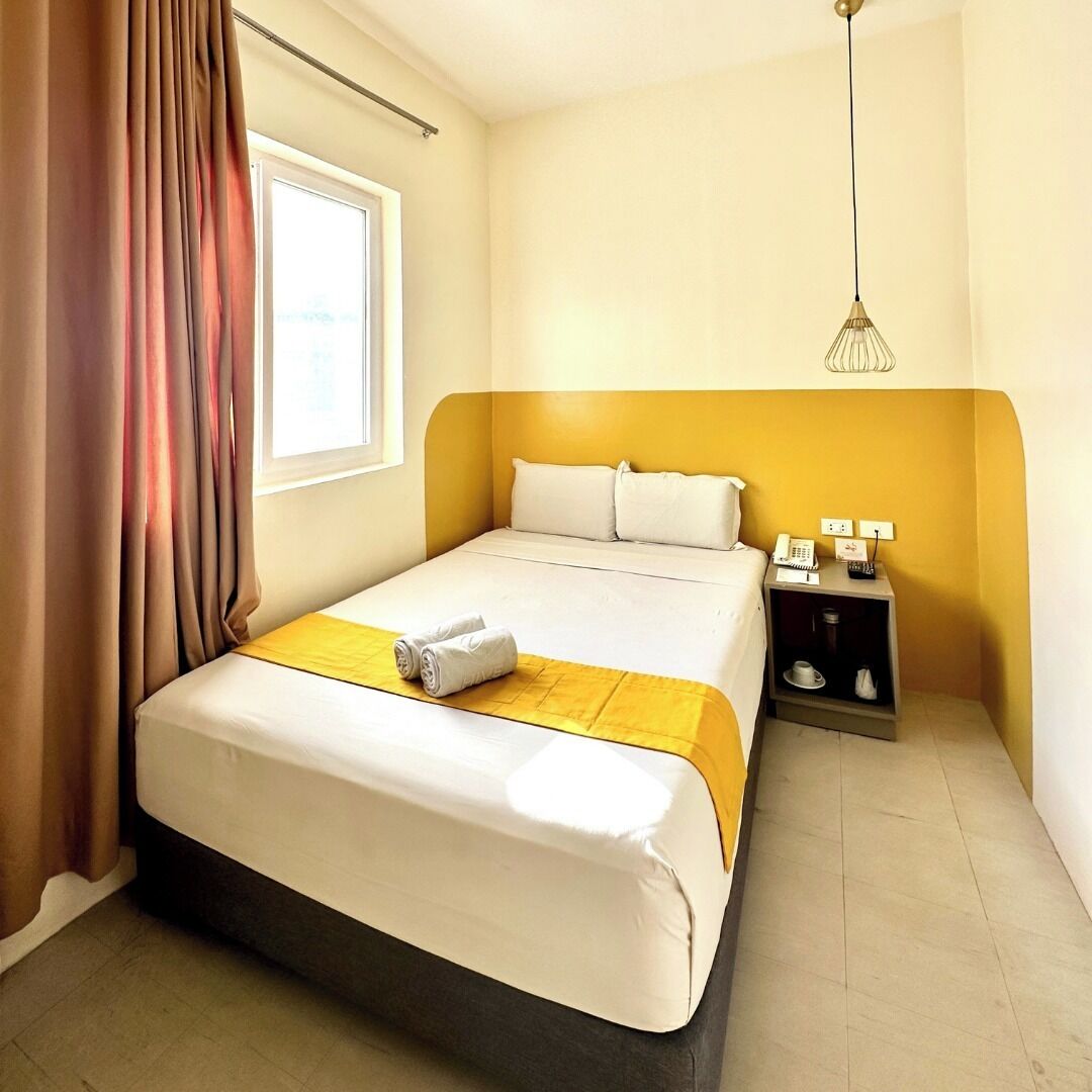 A hotel room with a bed, a bedside table with a lamp, and a window with curtains.