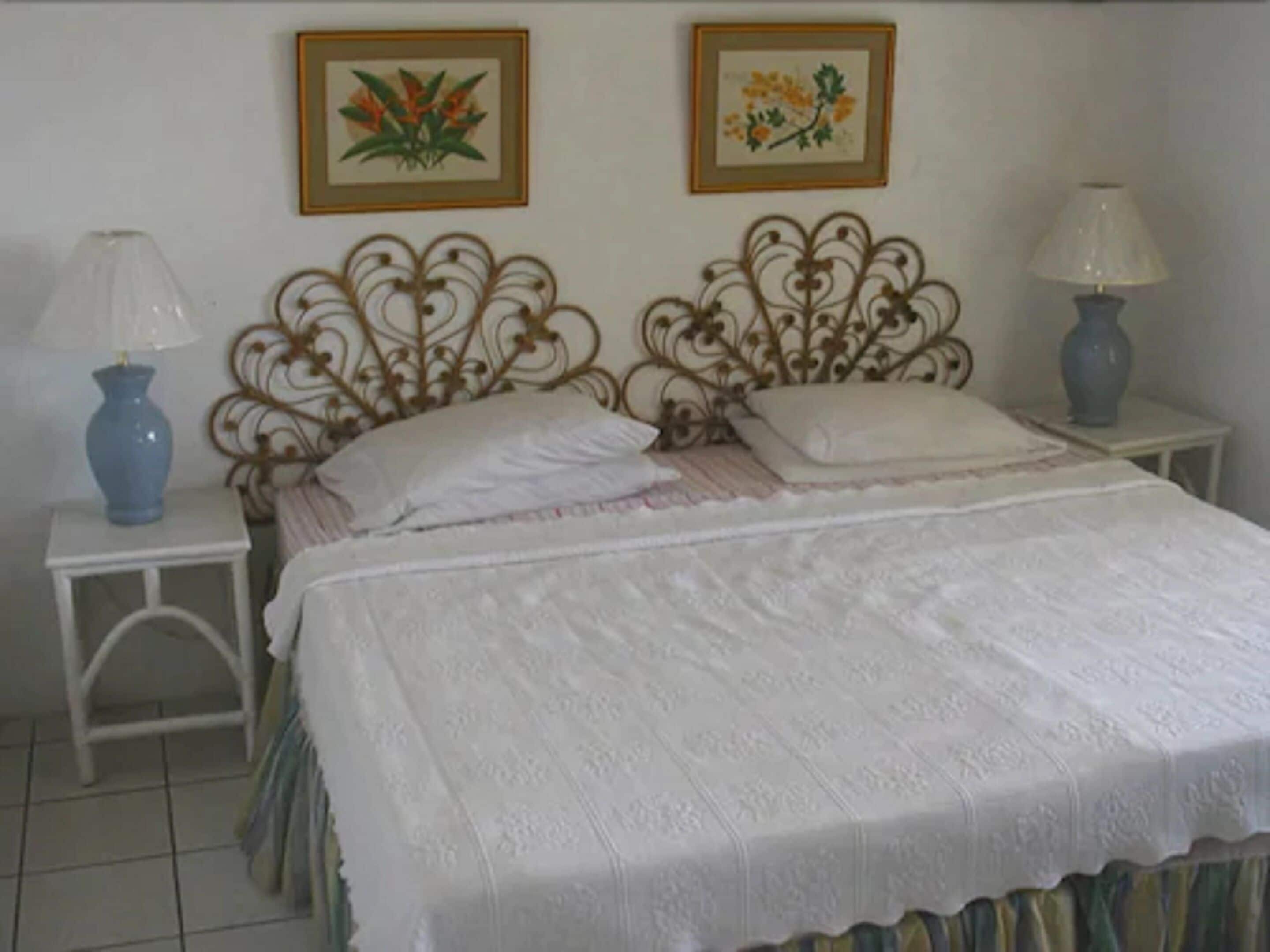 Una cama bien hecha con una colcha blanca y almohadas, flanqueada por dos mesitas de noche, una azul y otra gris.