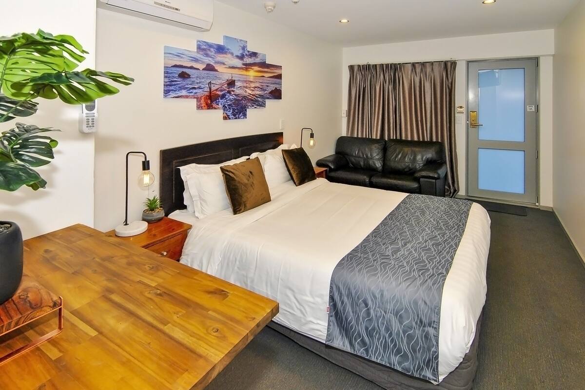 A hotel room with a bed, a wooden table, a leather sofa, a painting on the wall, and a window with curtains.