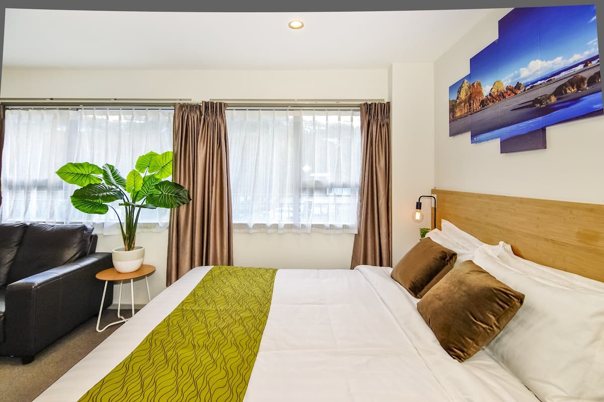 A hotel room with a bed, a potted plant, a sofa, and a view of the outdoors through the window.