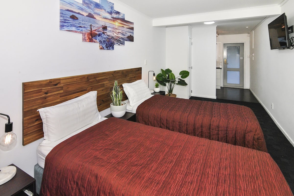 A hotel room with two beds, a wooden headboard, a potted plant, and a painting on the wall.