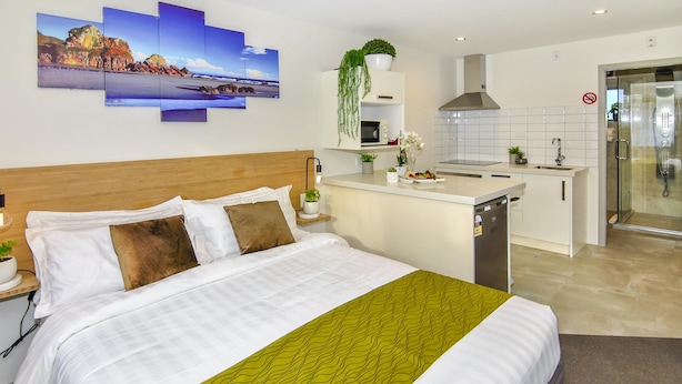 A hotel room with a bed, a bedside table with a lamp, a small kitchen area, and a shower. - Newlands Court Motel (Wellington)