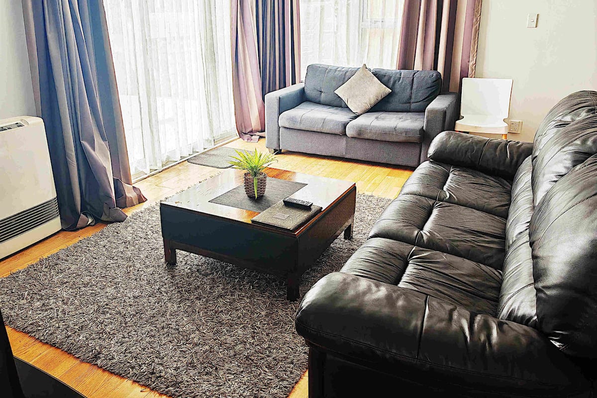 A living room with a grey sofa, a black leather sofa, a coffee table with a plant, and a large window with curtains.