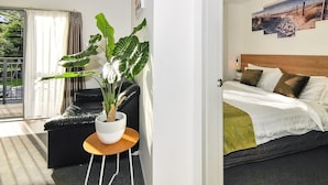 A hotel room with a bed, a black leather sofa, a potted plant, and a view of the outdoors. - Newlands Court Motel (Wellington)