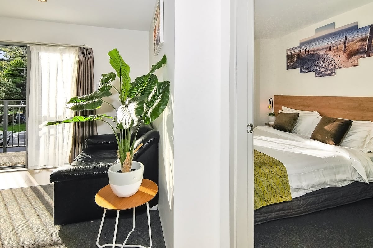 A hotel room with a bed, a black leather sofa, a potted plant, and a view of the outdoors.
