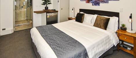 A hotel room with a large bed, a bedside table with a lamp, a small plant, and a wall-mounted artwork depicting a sunset over the ocean.