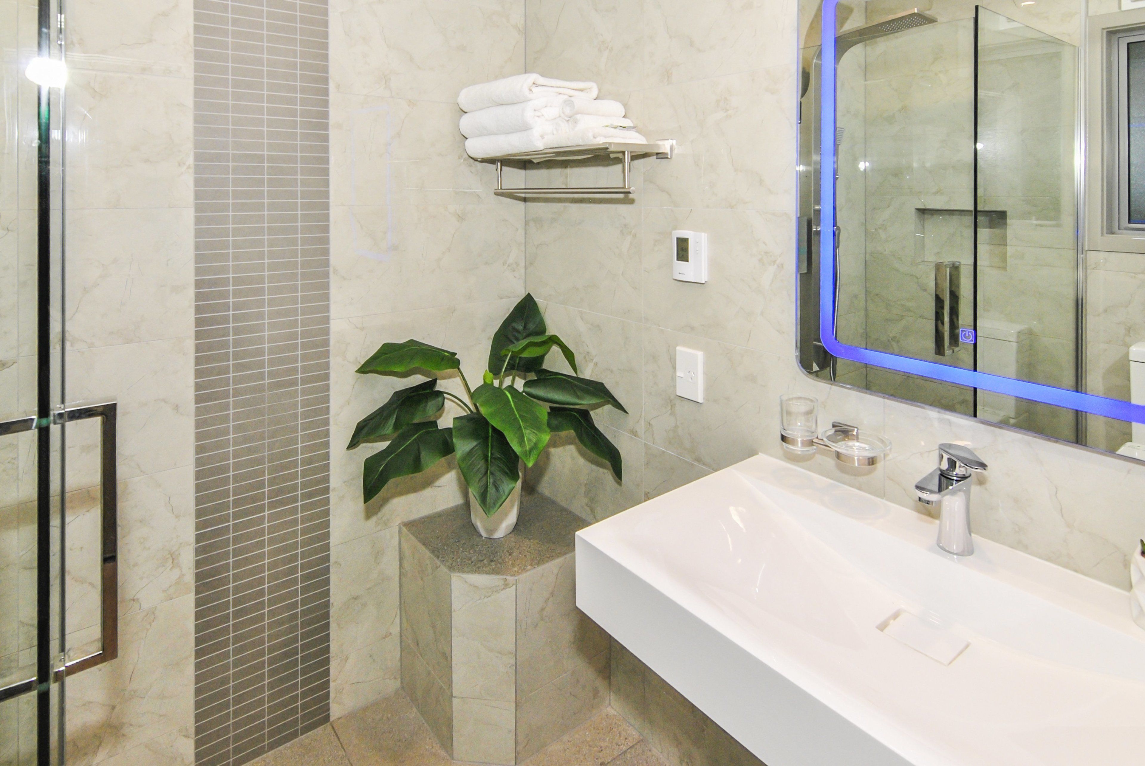 A modern bathroom with a large mirror, a white sink, a glass shower enclosure, and a potted plant.