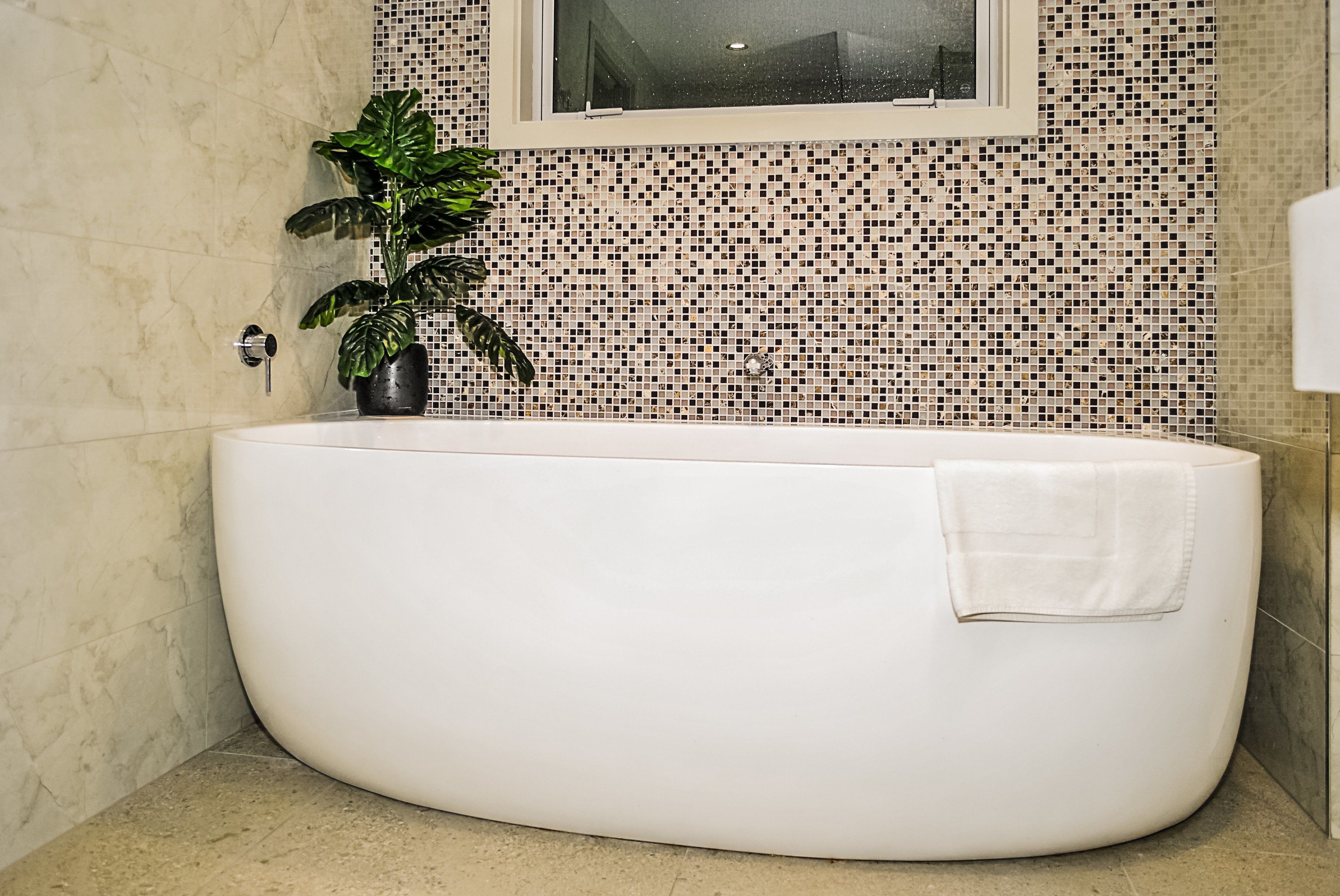 A modern bathroom with a freestanding bathtub, a potted plant, and a mosaic tile wall.