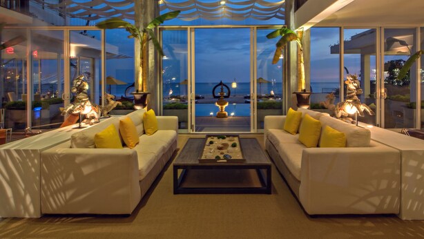 Un salon moderne avec des canapés blancs, des coussins jaunes et une table basse centrale. De grandes fenêtres offrent une vue sur la piscine et l’océan.