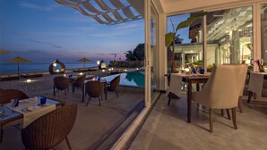 Un espace de restauration en plein air, avec des tables et des chaises, offrant une vue sur une piscine et une plage au crépuscule.