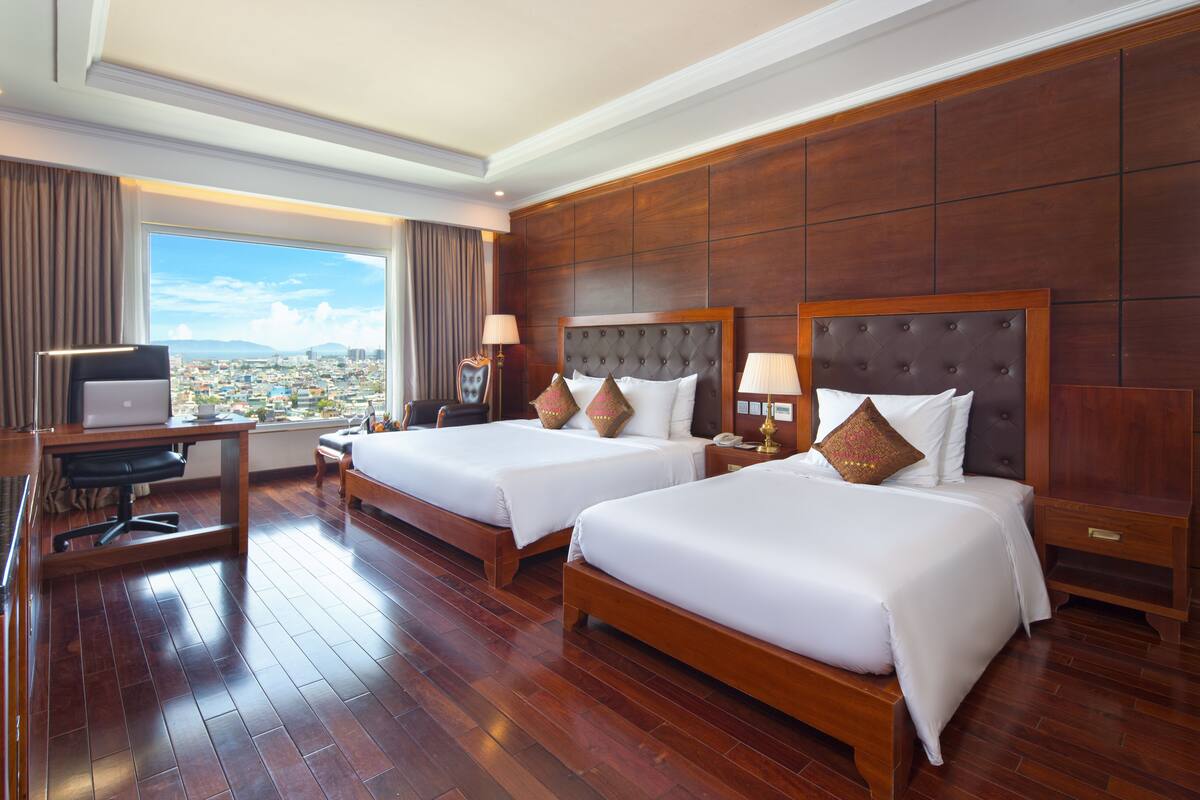 A hotel room with two beds, a desk with a laptop, and a view of the city.