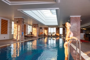 Ein Innenpool mit klaren Glasdecken, einem sonnendurchfluteten Innenraum und Liegestühlen am Pool.