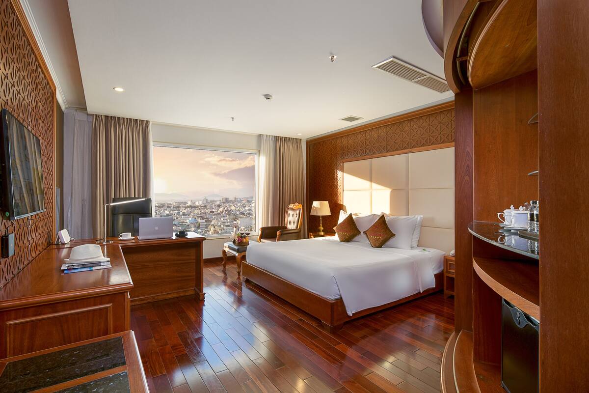 A hotel room with a large bed, a desk with a laptop, a television, and a view of the city.