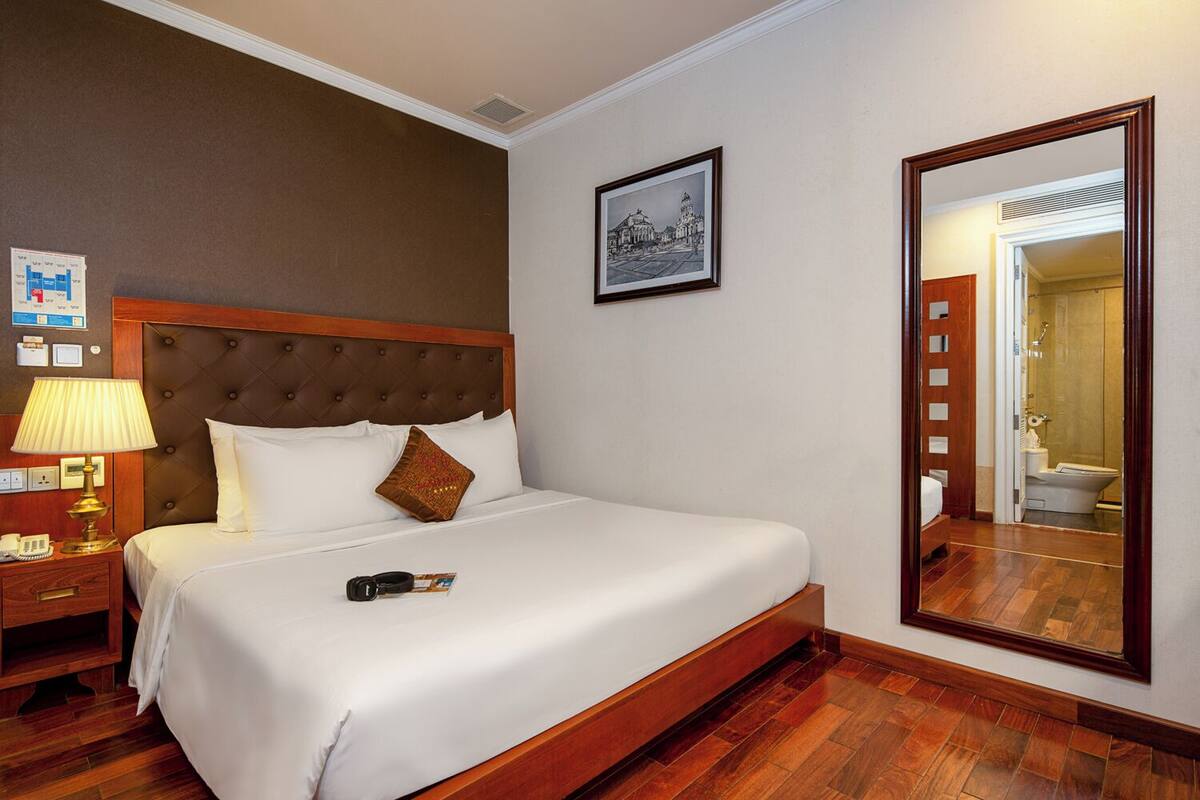 A hotel room with a large bed, a wooden headboard, a bedside table with a lamp, a framed picture on the wall, and a mirror reflecting the bathroom.