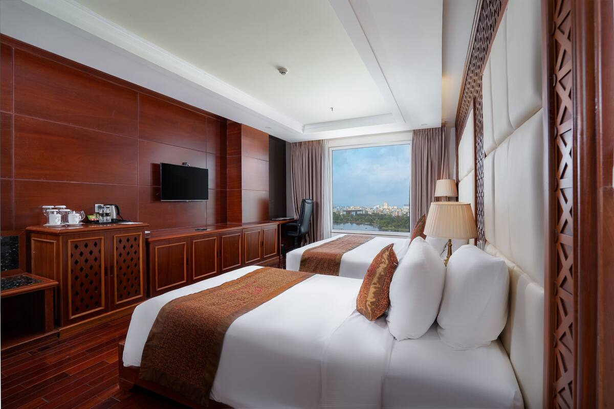 A hotel room with two beds, a TV, a desk with a coffee maker, and a view of a cityscape.