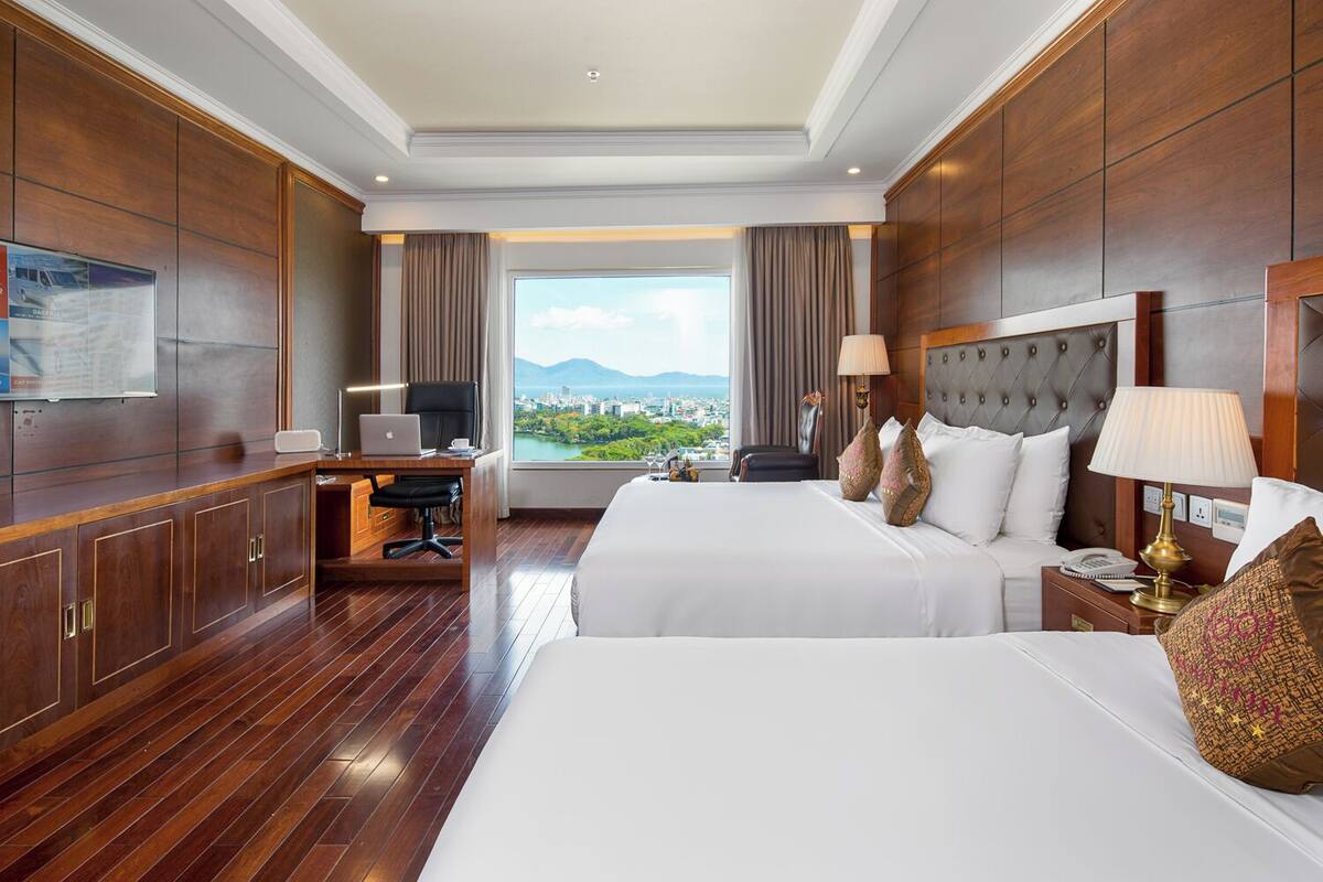 A hotel room with two beds, a desk with a laptop, a view of a cityscape, and a large window with curtains.