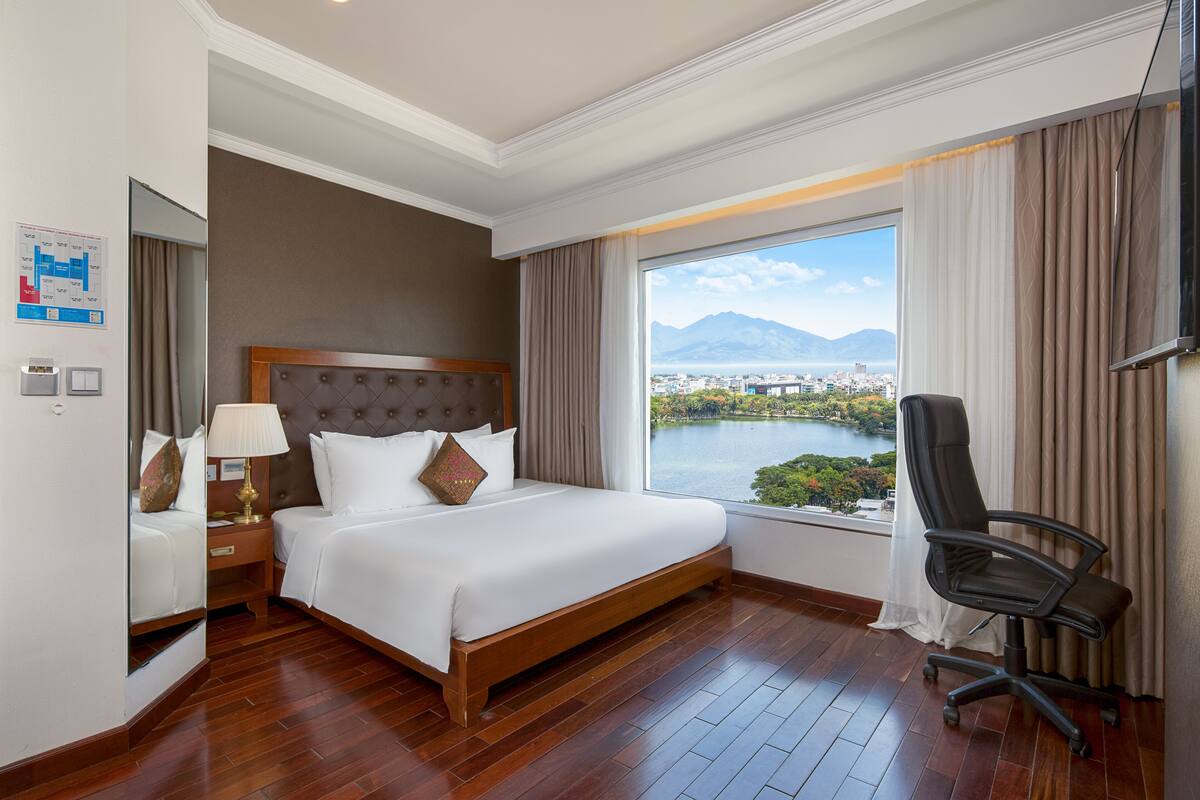 A hotel room with a large bed, a desk with a lamp, a chair, and a view of a lake and mountains.