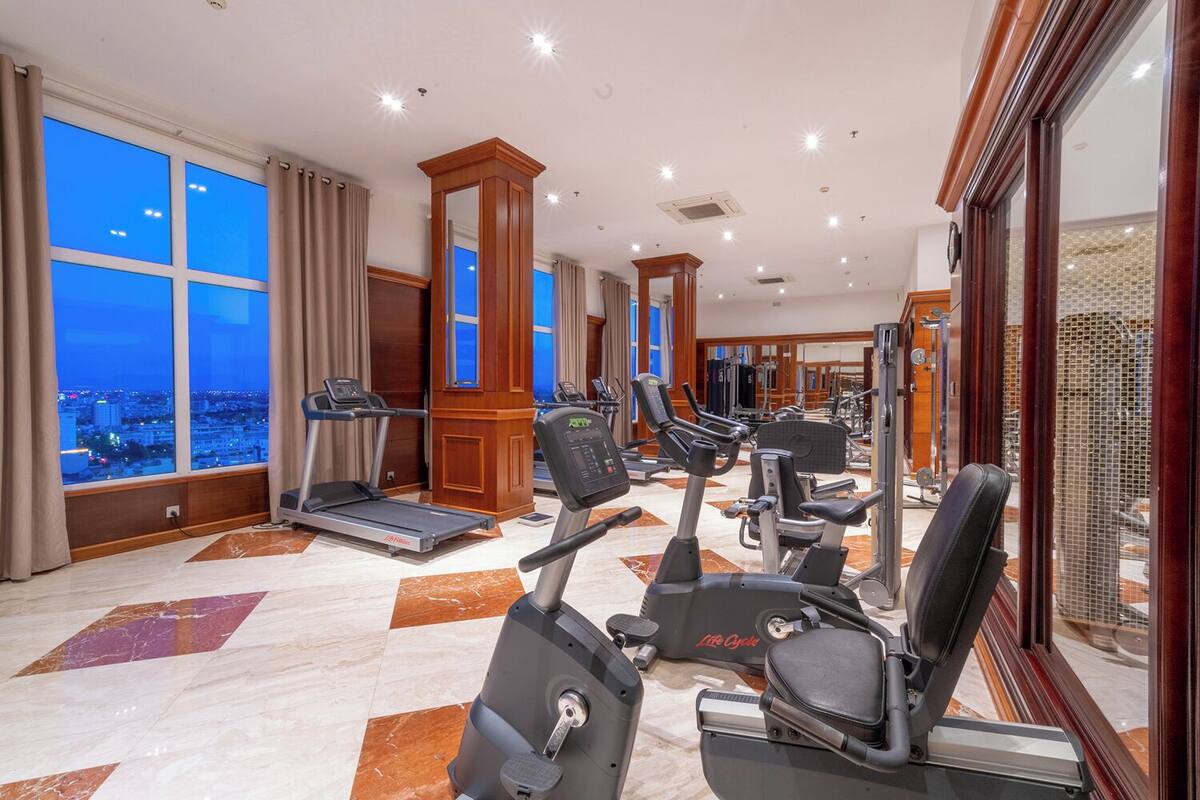 A hotel gym with treadmills, elliptical machines, and weight equipment, large windows with city views, and a marble floor.