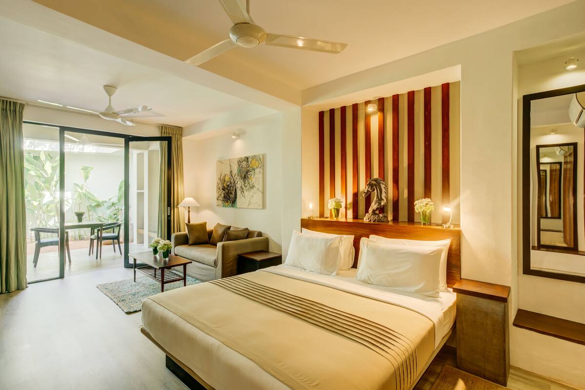 A hotel room with a large bed, a ceiling fan, a dining area with a table and chairs, and a sliding glass door leading to a balcony.