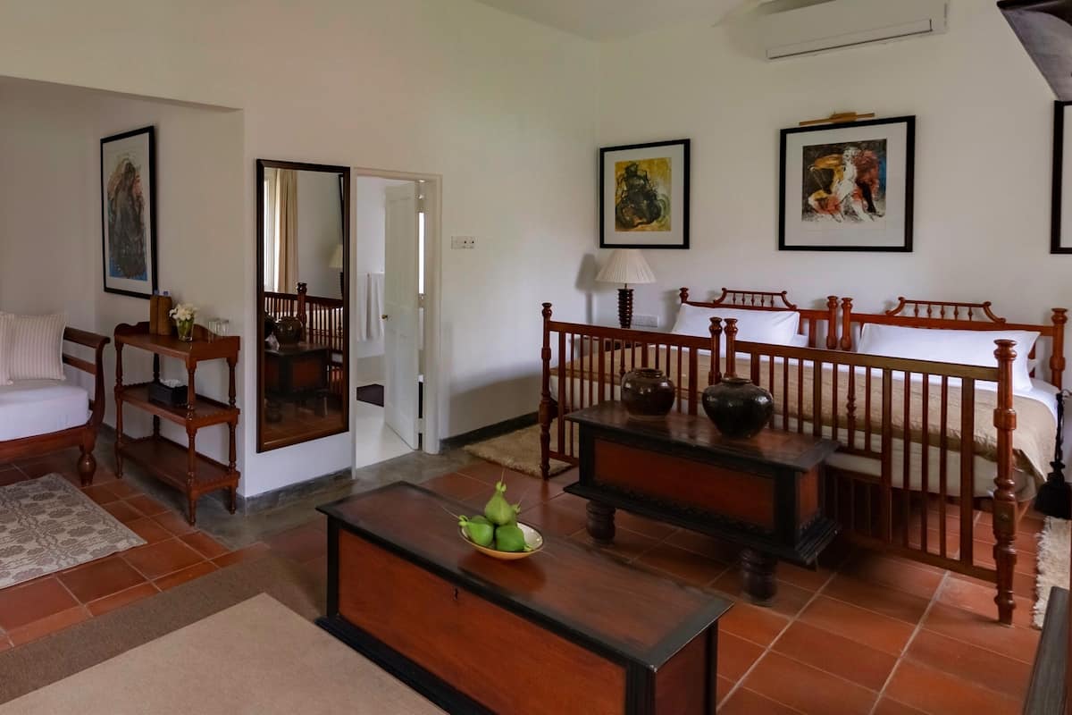 A living room with a sofa, coffee table, and a bed with a wooden headboard. There are framed pictures on the wall and a window with curtains.