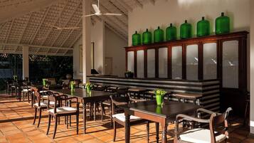 A spacious indoor dining area with wooden tables and chairs, a ceiling fan, and a cabinet with green bottles.