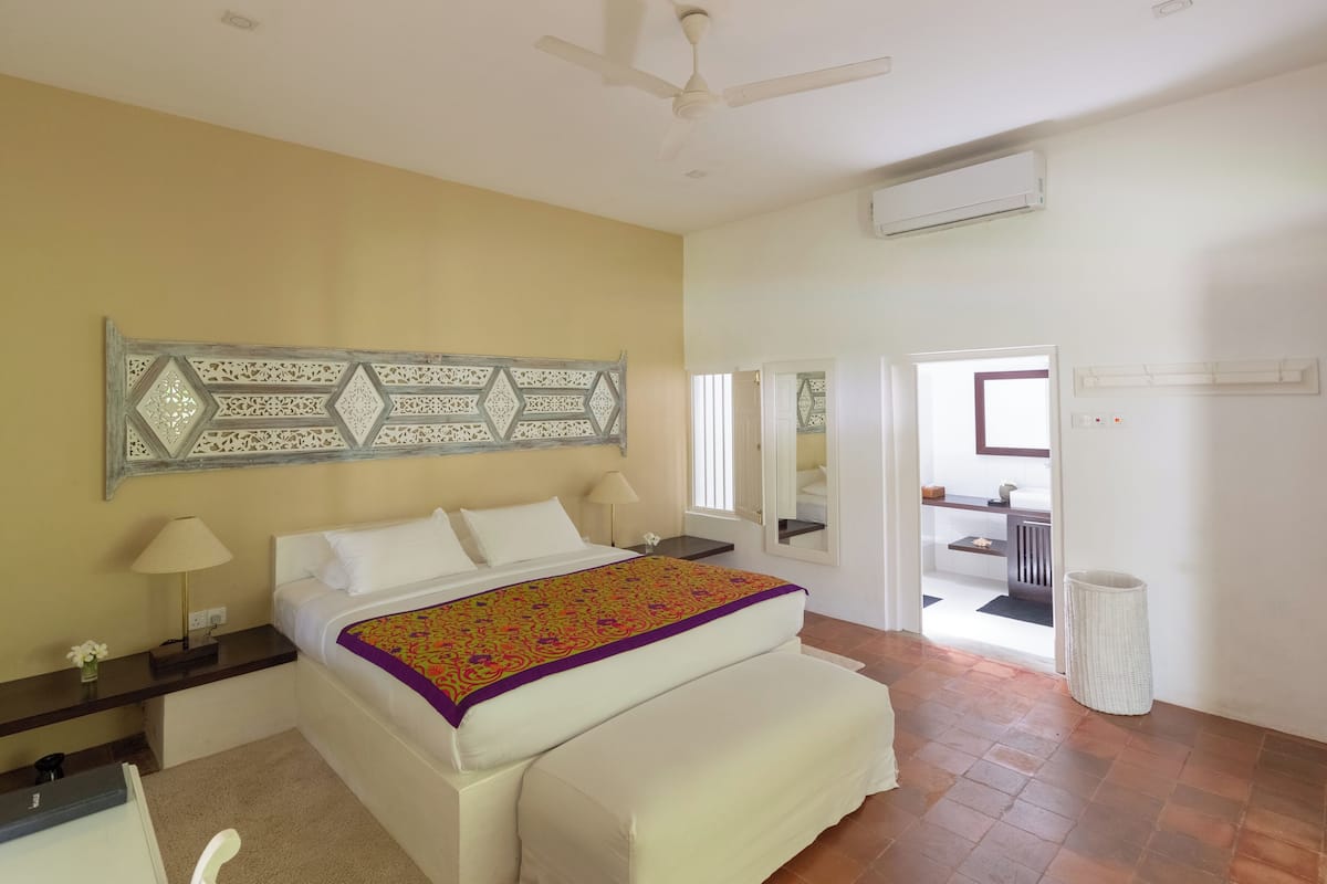 A hotel room with a bed, a nightstand, a lamp, a decorative wall piece, an air conditioning unit, and a view of the outside.