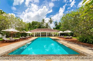 Une piscine rectangulaire, agrémentée de chaises longues et de parasols, entourée de verdure et d’un pavillon abrité.