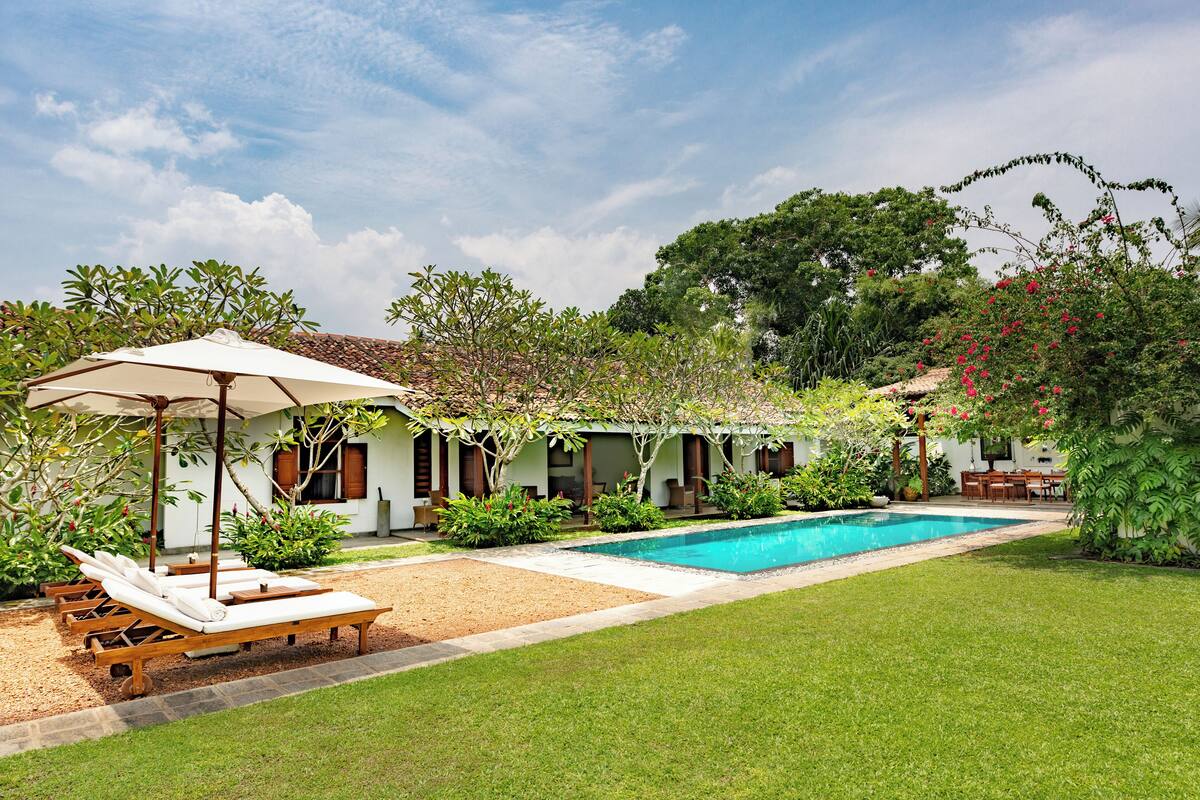 A poolside area with lounge chairs and a table under an umbrella, surrounded by greenery and a building with windows.