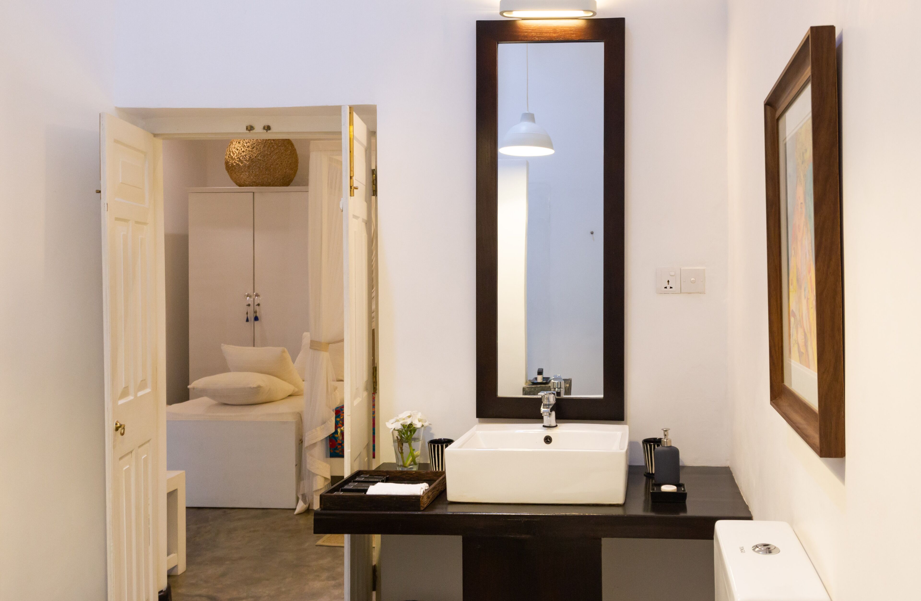 Un baño moderno con un lavamanos blanco, un espejo grande y una mesita de noche con lámpara.