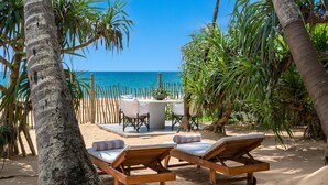 Un salon de plage doté de chaises longues en bois et d’une table avec des chaises, le tout entouré de palmiers et offrant une vue sur l’océan.