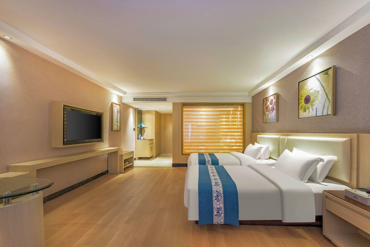 A hotel room with a large bed, a TV mounted on the wall, a bedside table, and a window with blinds.