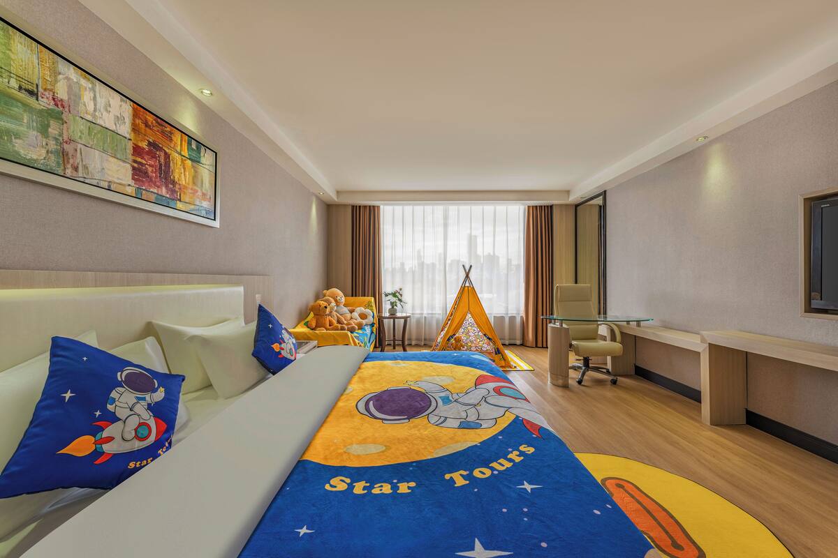 A hotel room with a bed featuring a space-themed bedspread, a desk with a chair, a window with curtains, and a wall-mounted TV.