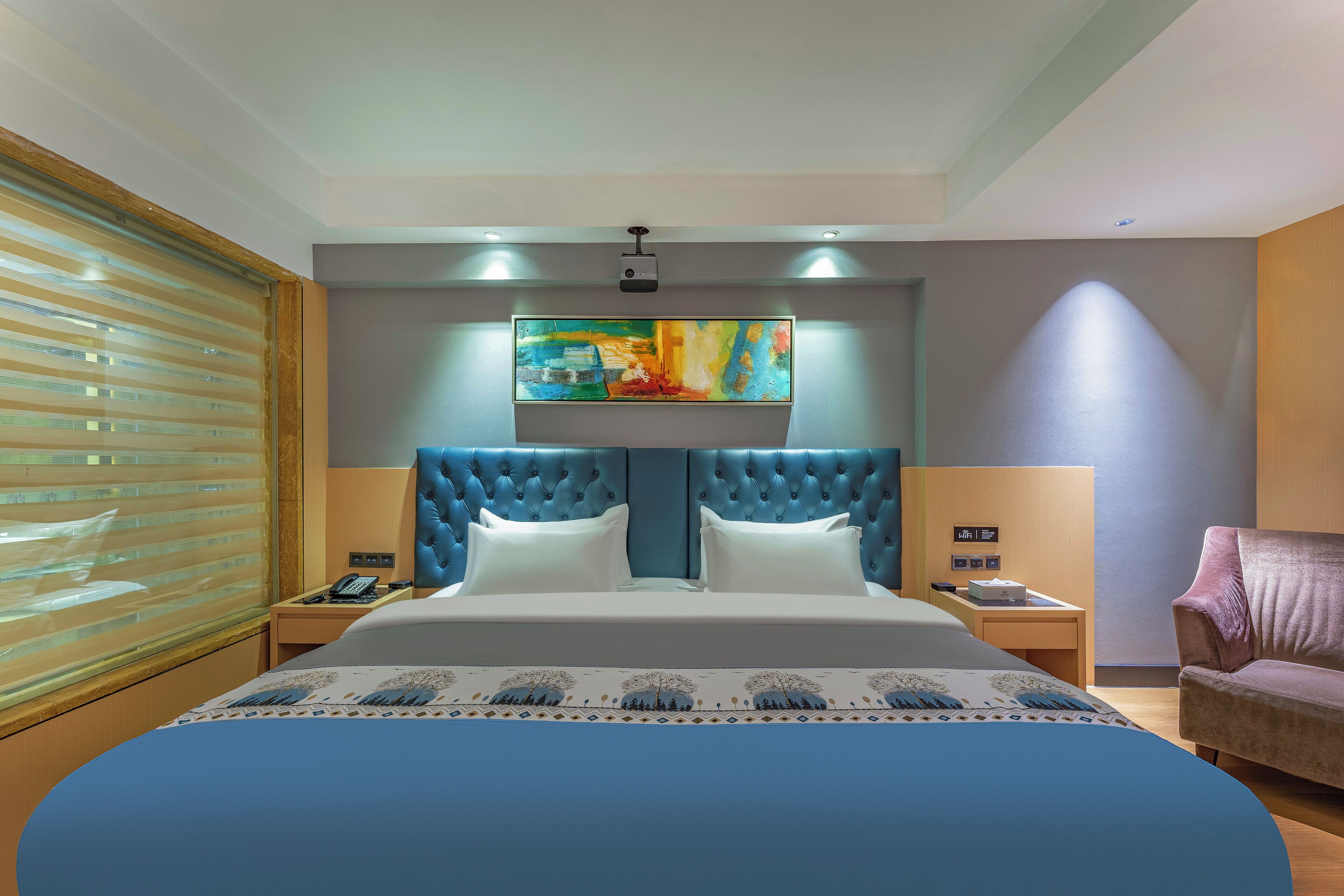 Une chambre d’hôtel avec un grand lit, une tête de lit capitonnée, un tableau au-dessus du lit, une table de chevet avec un téléphone et une fenêtre avec des stores.